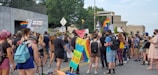 A group of people participating in a street protest, holding various signs and banners supporting Black Lives Matter and LGBTQ+ rights. Many of the protesters are wearing backpacks and casual clothing, and some are masked. Rainbow flags and other placards are visible in the crowd, indicating a call for justice and equality.