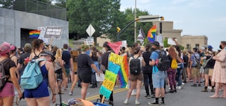 A group of people participating in a street protest, holding various signs and banners supporting Black Lives Matter and LGBTQ+ rights. Many of the protesters are wearing backpacks and casual clothing, and some are masked. Rainbow flags and other placards are visible in the crowd, indicating a call for justice and equality.