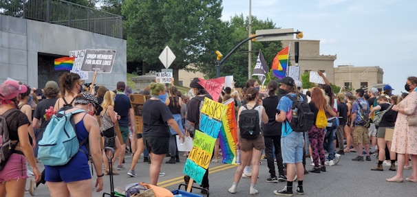 A group of people participating in a street protest, holding various signs and banners supporting Black Lives Matter and LGBTQ+ rights. Many of the protesters are wearing backpacks and casual clothing, and some are masked. Rainbow flags and other placards are visible in the crowd, indicating a call for justice and equality.