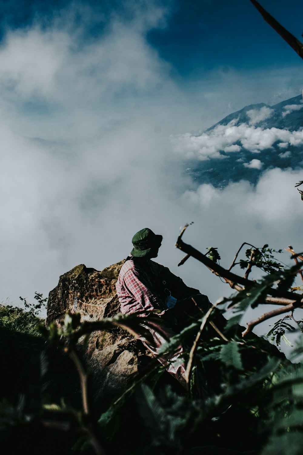 man in red and black jacket sitting on rock mountain under white clouds during daytime