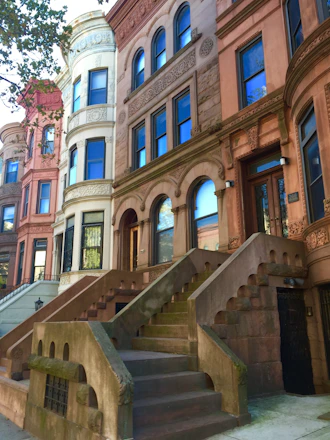 A row of historic, multi-story brownstone buildings with detailed architectural embellishments. The buildings feature arched windows and doorways, elaborate stone carvings, and large staircases leading up to the main entrances. Trees with green leaves are visible to the left side, adding a natural element to the urban setting.