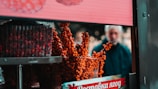 A stall displays vibrant orange berries on branches, with plastic containers of small round fruits stacked nearby. A blurred figure stands in the background, adding depth to the scene. The background sign features Cyrillic writing.