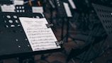 A music stand holds a sheet of music in focus while additional music stands and chairs are visible out of focus in the background. The setting appears to be an orchestral or ensemble environment, with several empty chairs suggesting preparation for a performance.