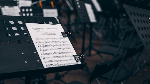 A music stand holds a sheet of music in focus while additional music stands and chairs are visible out of focus in the background. The setting appears to be an orchestral or ensemble environment, with several empty chairs suggesting preparation for a performance.