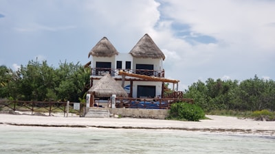A two-story house with a thatched roof sits on a sandy beach. The front of the house features wooden railings and balconies. The surrounding area includes lush greenery and the edges of an ocean or large body of water nearby.