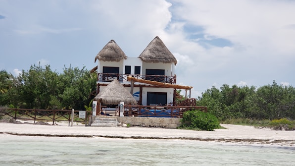 A two-story house with a thatched roof sits on a sandy beach. The front of the house features wooden railings and balconies. The surrounding area includes lush greenery and the edges of an ocean or large body of water nearby.