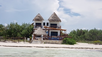A two-story house with a thatched roof sits on a sandy beach. The front of the house features wooden railings and balconies. The surrounding area includes lush greenery and the edges of an ocean or large body of water nearby.
