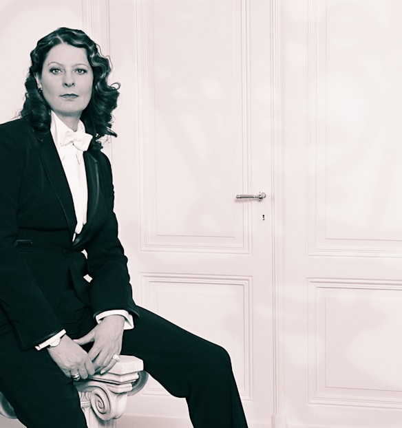 A person in a formal black suit and white bow tie is seated indoors against a plain, light-colored panel door. The person has wavy hair and is gazing directly at the camera, exuding a poised and confident demeanor.