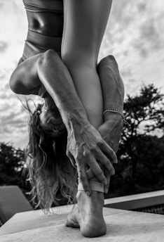 A person performing a yoga pose outdoors, bending forward and holding their foot with both hands. The focus is on their muscular arms and legs, with an emphasis on body flexibility and strength. The background shows cloudy skies and trees in a natural setting.