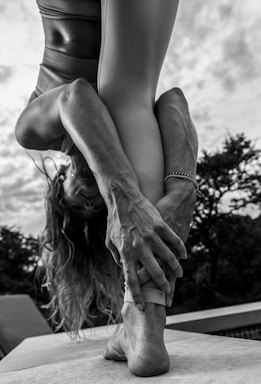 A person performing a yoga pose outdoors, bending forward and holding their foot with both hands. The focus is on their muscular arms and legs, with an emphasis on body flexibility and strength. The background shows cloudy skies and trees in a natural setting.
