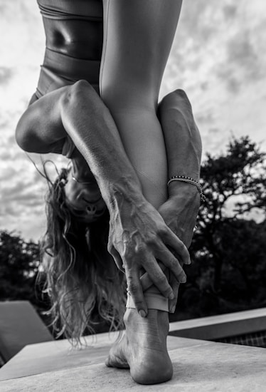 A person performing a yoga pose outdoors, bending forward and holding their foot with both hands. The focus is on their muscular arms and legs, with an emphasis on body flexibility and strength. The background shows cloudy skies and trees in a natural setting.