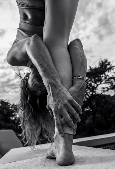 A person performing a yoga pose outdoors, bending forward and holding their foot with both hands. The focus is on their muscular arms and legs, with an emphasis on body flexibility and strength. The background shows cloudy skies and trees in a natural setting.