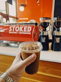 A hand holds a cold brew coffee in a clear plastic cup with a lid and ice visible inside. In the background, a red espresso machine with the words 'STOKED Roasters + Coffeehouse' is seen, along with two black coffee grinders filled with coffee beans. The setting has a warm, inviting interior with wood and orange tones.