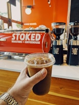 A hand holds a cold brew coffee in a clear plastic cup with a lid and ice visible inside. In the background, a red espresso machine with the words 'STOKED Roasters + Coffeehouse' is seen, along with two black coffee grinders filled with coffee beans. The setting has a warm, inviting interior with wood and orange tones.
