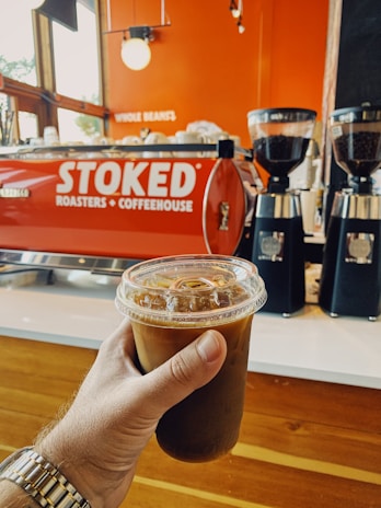 A hand holds a cold brew coffee in a clear plastic cup with a lid and ice visible inside. In the background, a red espresso machine with the words 'STOKED Roasters + Coffeehouse' is seen, along with two black coffee grinders filled with coffee beans. The setting has a warm, inviting interior with wood and orange tones.