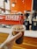 A hand holds a cold brew coffee in a clear plastic cup with a lid and ice visible inside. In the background, a red espresso machine with the words 'STOKED Roasters + Coffeehouse' is seen, along with two black coffee grinders filled with coffee beans. The setting has a warm, inviting interior with wood and orange tones.