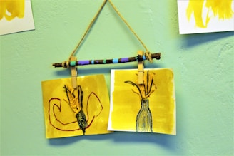 Two yellow-toned artworks are attached to a painted stick with clothespins, hanging against a light green textured wall. The left drawing features abstract shapes in red and black, while the right illustrates a vase with a textured design, holding branches or flowers.
