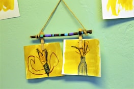 Two yellow-toned artworks are attached to a painted stick with clothespins, hanging against a light green textured wall. The left drawing features abstract shapes in red and black, while the right illustrates a vase with a textured design, holding branches or flowers.
