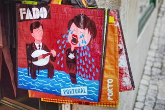 A colorful illustration on a shopping bag depicts two stylized figures, one playing a string instrument and the other singing with exaggerated tears flowing. The background features wording like 'FADO' and 'PORTUGAL' with a wavy blue pattern at the bottom representing water. The scene suggests a musical theme, likely around traditional Portuguese music, and the emotion is captured through the expressive, cartoon-like drawing style.