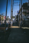 A busy urban street scene featuring a sidewalk lined with utility poles and overhead wires. A row of parked cars is visible on the left side, with several buildings and storefronts lining the other side. A palm tree can be seen in the background, along with various billboards and signs.