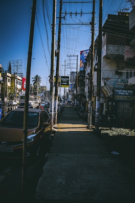 A busy urban street scene featuring a sidewalk lined with utility poles and overhead wires. A row of parked cars is visible on the left side, with several buildings and storefronts lining the other side. A palm tree can be seen in the background, along with various billboards and signs.