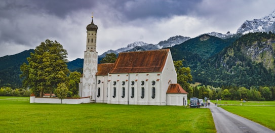 A picturesque scene of a white church with a tall spire situated in a lush green field, surrounded by trees. In the background, the landscape features mountain ranges with snow-capped peaks under a cloudy sky. A narrow road leads towards the church with a few people walking nearby.