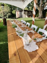 A long wooden table is set outdoors with a simple, elegant arrangement for an event. Glasses are turned upside down on napkins, with green foliage and candles as centerpieces. White chairs surround the table, all under a canopy. A garden with a small rustic shed and trees provides a serene backdrop.