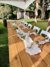 A long wooden table is set outdoors with a simple, elegant arrangement for an event. Glasses are turned upside down on napkins, with green foliage and candles as centerpieces. White chairs surround the table, all under a canopy. A garden with a small rustic shed and trees provides a serene backdrop.