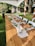 A long wooden table is set outdoors with a simple, elegant arrangement for an event. Glasses are turned upside down on napkins, with green foliage and candles as centerpieces. White chairs surround the table, all under a canopy. A garden with a small rustic shed and trees provides a serene backdrop.