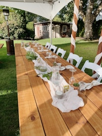 A long wooden table is set outdoors with a simple, elegant arrangement for an event. Glasses are turned upside down on napkins, with green foliage and candles as centerpieces. White chairs surround the table, all under a canopy. A garden with a small rustic shed and trees provides a serene backdrop.