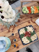 A rustic wooden table adorned with a variety of foods, including a three-tiered white cake decorated with flowers and greenery. A platter of cubed cheeses and sausages is arranged with kale. Adjacent to this is a tray of fresh vegetables like broccoli, cherry tomatoes, cucumber slices, and carrot sticks. Nearby, bottles of dressing are positioned next to a large, clear glass container possibly filled with a beverage on ice.