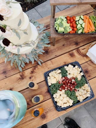 A rustic wooden table adorned with a variety of foods, including a three-tiered white cake decorated with flowers and greenery. A platter of cubed cheeses and sausages is arranged with kale. Adjacent to this is a tray of fresh vegetables like broccoli, cherry tomatoes, cucumber slices, and carrot sticks. Nearby, bottles of dressing are positioned next to a large, clear glass container possibly filled with a beverage on ice.