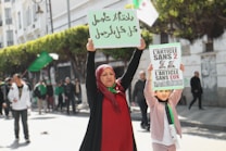 A woman and a child participate in a public demonstration, each holding up signs with political messages. The woman wears a red headscarf and the child is dressed in light-colored clothing. They stand in a street lined with trees and other people are visible in the background, some holding flags.