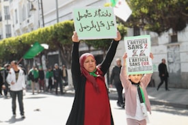 A woman and a child participate in a public demonstration, each holding up signs with political messages. The woman wears a red headscarf and the child is dressed in light-colored clothing. They stand in a street lined with trees and other people are visible in the background, some holding flags.