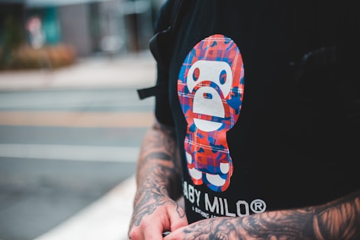 A person with tattooed arms is wearing a black T-shirt featuring a colorful graphic of a stylized character. The graphic consists of red, blue, and white colors arranged in a checkered pattern. The visible background is blurred, indicating an urban environment.