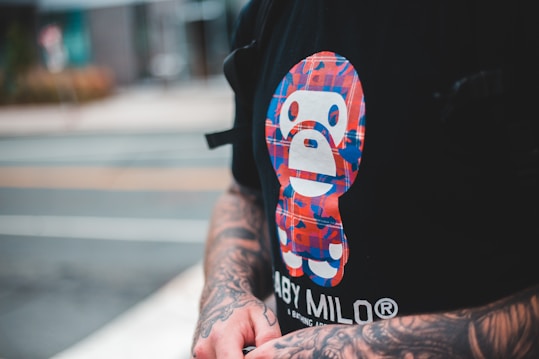 A person with tattooed arms is wearing a black T-shirt featuring a colorful graphic of a stylized character. The graphic consists of red, blue, and white colors arranged in a checkered pattern. The visible background is blurred, indicating an urban environment.