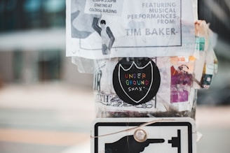 A pole is covered with various posters and stickers, including a black circular sticker with colorful text reading 'UNDERGROUND SNAX.' The posters appear weathered, with some torn edges and overlapping layers. The background is blurred, indicating an urban setting.