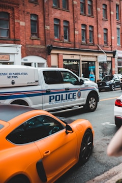 A city street scene featuring a police vehicle from the Royal Newfoundland Constabulary parked alongside a bright orange sports car. The background consists of a row of brick buildings with various shops and businesses.