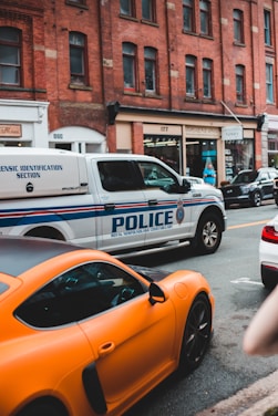 A city street scene featuring a police vehicle from the Royal Newfoundland Constabulary parked alongside a bright orange sports car. The background consists of a row of brick buildings with various shops and businesses.