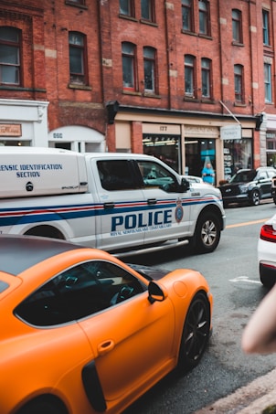 A city street scene featuring a police vehicle from the Royal Newfoundland Constabulary parked alongside a bright orange sports car. The background consists of a row of brick buildings with various shops and businesses.