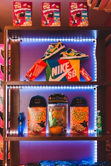 A shelf filled with various branded products, including colorful cereal boxes on the top shelf labeled as Froot Loops with Marshmallows. The middle shelf features a pair of multicolored sneakers positioned above a Nike SB shoebox, surrounded by red boxes labeled Supreme and Ziploc. The bottom shelf displays two caps, multiple boxes of Reese's Puffs cereal, a small bowl, and a blue Pepsi bottle to the left with a green 7up bottle to the right. The entire display is illuminated with LED lights, creating a vibrant and energetic atmosphere.