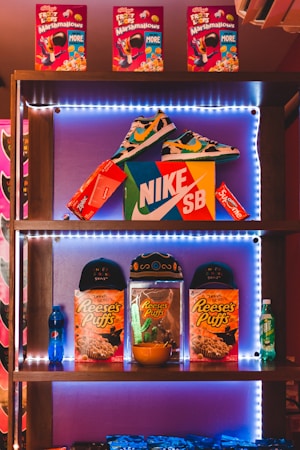 A shelf filled with various branded products, including colorful cereal boxes on the top shelf labeled as Froot Loops with Marshmallows. The middle shelf features a pair of multicolored sneakers positioned above a Nike SB shoebox, surrounded by red boxes labeled Supreme and Ziploc. The bottom shelf displays two caps, multiple boxes of Reese's Puffs cereal, a small bowl, and a blue Pepsi bottle to the left with a green 7up bottle to the right. The entire display is illuminated with LED lights, creating a vibrant and energetic atmosphere.
