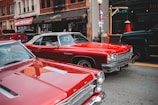 Two classic red cars are parked on a city street in front of a clothing store with a sign reading 'Luxurious Outerwear'. Several people can be seen in the background along the sidewalk. The scene includes other parked vehicles, a lamp post, and a visible shop window.