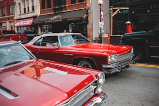 Two classic red cars are parked on a city street in front of a clothing store with a sign reading 'Luxurious Outerwear'. Several people can be seen in the background along the sidewalk. The scene includes other parked vehicles, a lamp post, and a visible shop window.