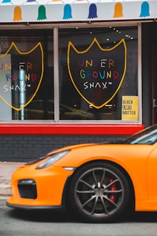 An orange sports car is parked in front of a shop. The shop has large windows with a sign displaying the words 'UNDER GROUND SNAX' in colorful letters inside shield-like outlines. Above the windows, a canopy with multicolored decorative shapes is visible. A yellow sign with 'BLACK LIVES MATTER' is placed near the entrance.