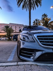 A silver luxury car is parked in an open lot with palm trees in the background. The facade of a light pink building with balconies is visible, and the scene captures the transition from day to night with a blue sky. The car's front is prominent, highlighting its shiny grille and headlights.