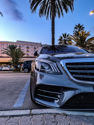 A silver luxury car is parked in an open lot with palm trees in the background. The facade of a light pink building with balconies is visible, and the scene captures the transition from day to night with a blue sky. The car's front is prominent, highlighting its shiny grille and headlights.