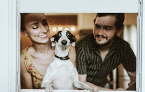 A woman and a man are smiling and looking affectionately at a black and white dog. The dog is positioned between them, framed by a window, and appears to be looking directly at the camera. Soft lighting creates a warm and intimate atmosphere in the room.