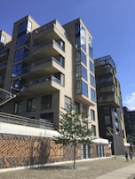 A modern multi-story residential building with numerous balconies and large glass windows. The building features a mix of light beige and dark panels, with a decorative brick wall pattern near the base. In the foreground, a small tree is planted on a cobblestone path, and two people are walking along the sidewalk. The sky is clear and blue, contributing to a bright atmosphere.