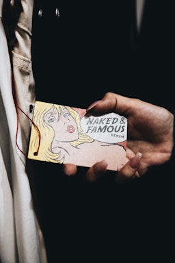 A hand is holding a vintage-style tag featuring a pop art illustration of a blonde woman alongside text that reads 'Naked & Famous Denim'. The background is dark, and the focus is on the tag.